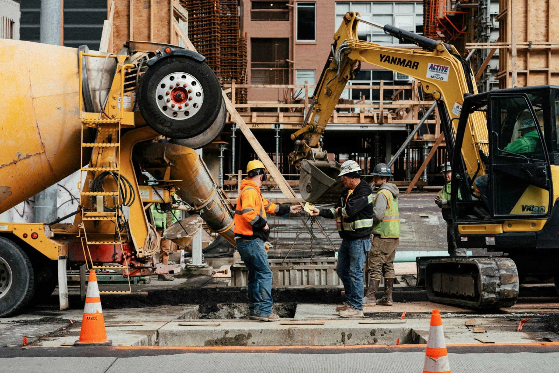 Two construction workers in hard hats exchanging a delivery slip on a site between a mixer truck and an excavator
